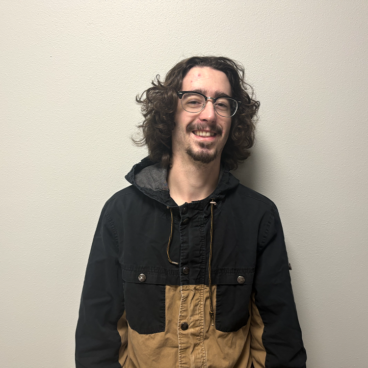 Young student with dark brown near shoulder length curly hair and a half-black half brown jacket.