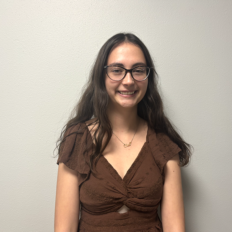 Young woman with brown v-neck shirt, long brown hair , and cat-eye glasses smiling.