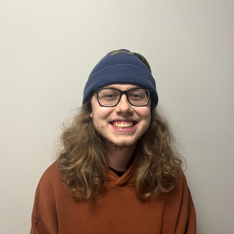 Young student with shoulder-length curly light brown hair, blue beanie and a orange-brown sweatshirt smiling.